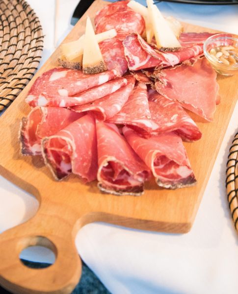 Tagliere di legno con assortimento di salumi affettati, formaggi stagionati e una ciotolina di miele con pinoli.