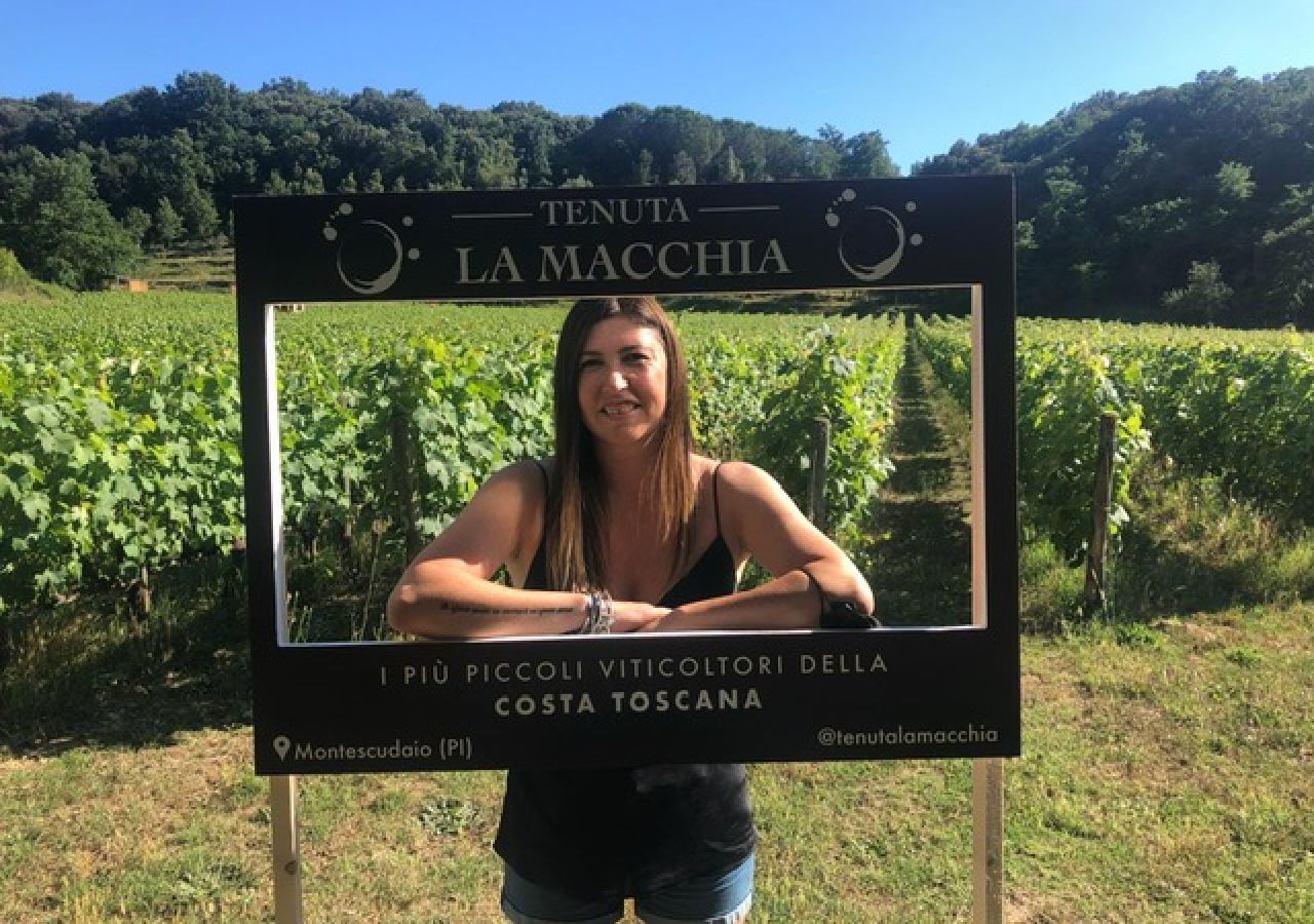 Una donna posa sorridente tra i filari della Tenuta La Macchia, affacciata all’interno di una cornice con il nome dell’azienda e la scritta “I più piccoli viticoltori della Costa Toscana”. Sullo sfondo si vedono il vigneto, la vegetazione e il cielo azzurro.