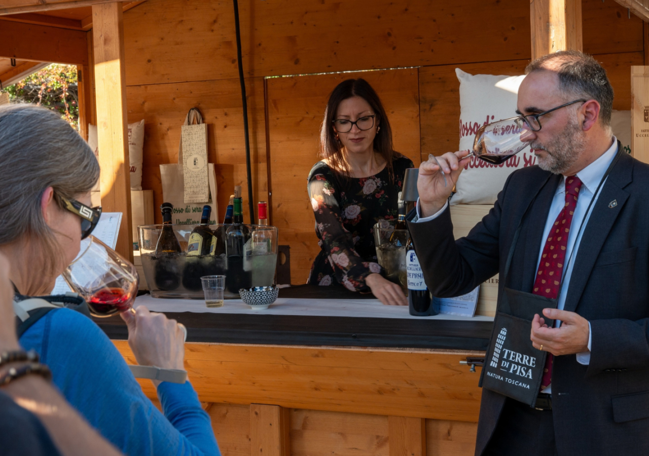 Sommelier guida una degustazione di vino davanti allo stand di una cantina durante il Terre di Pisa Food & Wine Festival, mentre i partecipanti osservano e assaggiano i vini proposti.