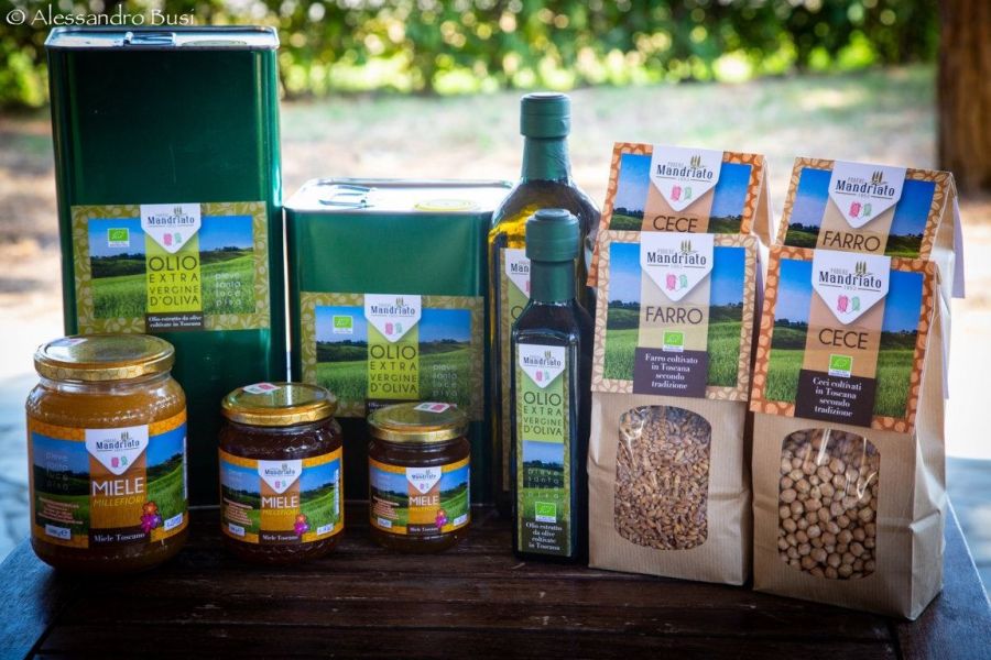 Selezione di prodotti biologici di Podere Mandriato, con olio extravergine di oliva, miele millefiori, farro e ceci confezionati, disposti su un tavolo all’aperto.