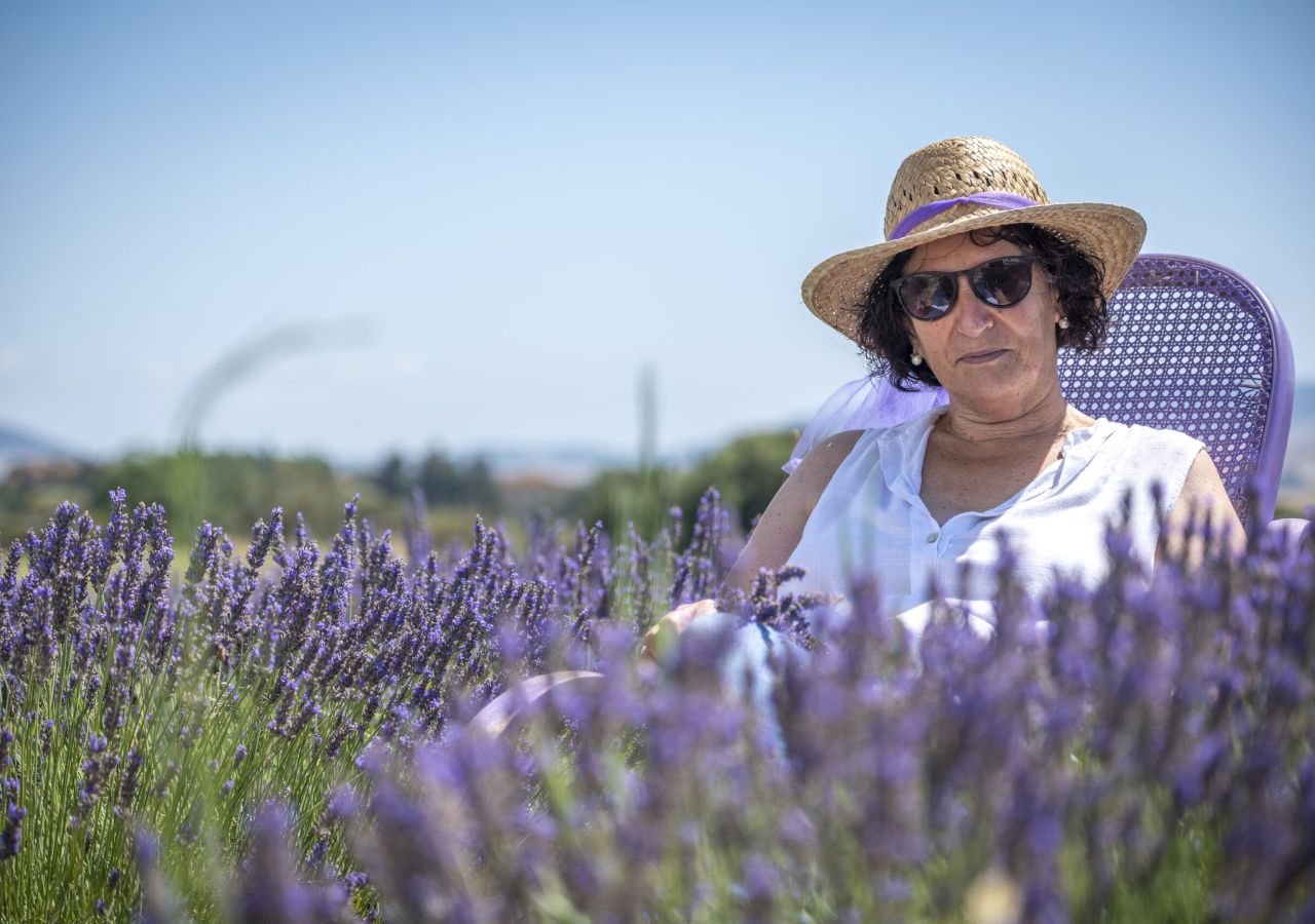 Una donna con cappello di paglia e occhiali da sole siede su una poltrona viola in mezzo a un campo di lavanda, sotto un cielo limpido.