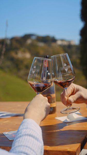 Due persone brindano con calici di vino rosato durante una degustazione all’aperto; sullo sfondo, sfocati, si intravedono le colline e una bottiglia di Castelvecchio.