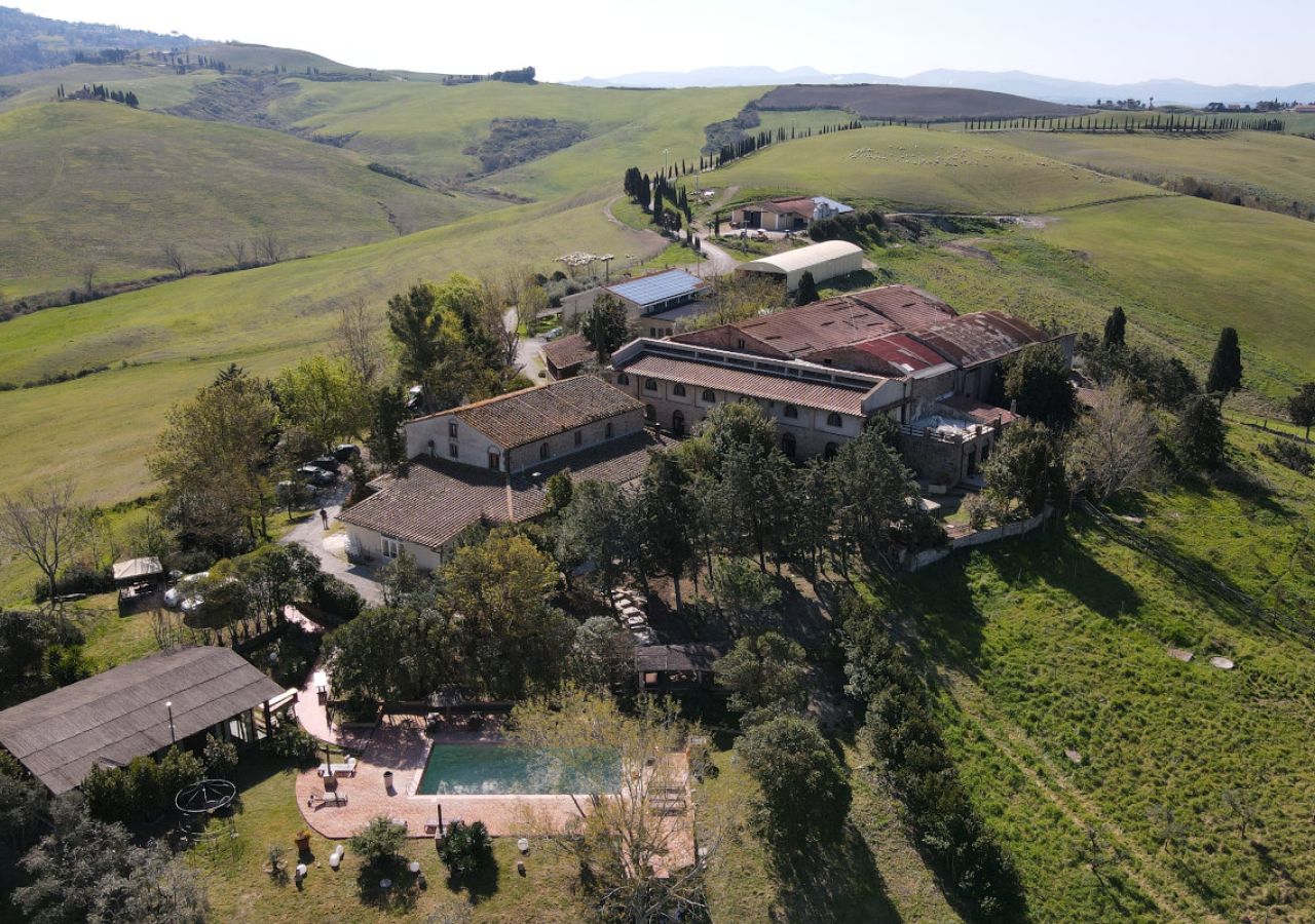 Veduta aerea della Fattoria Lischeto, immersa tra le colline toscane, con gli edifici rurali, la piscina e i filari di cipressi.