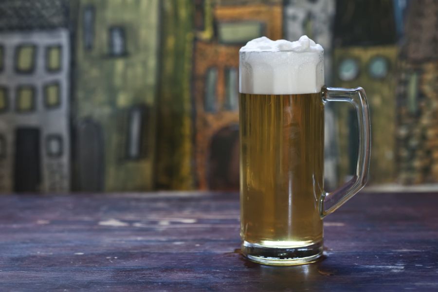 Boccale di birra artigianale chiara con abbondante schiuma bianca, appoggiato su un tavolo con sfondo pittorico sfocato.