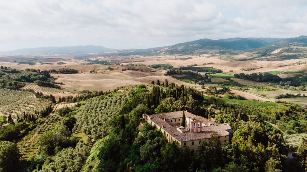 Veduta aerea della Badia di Morrona, circondata da cipressi, oliveti e vigneti, con le colline toscane che si estendono sullo sfondo.