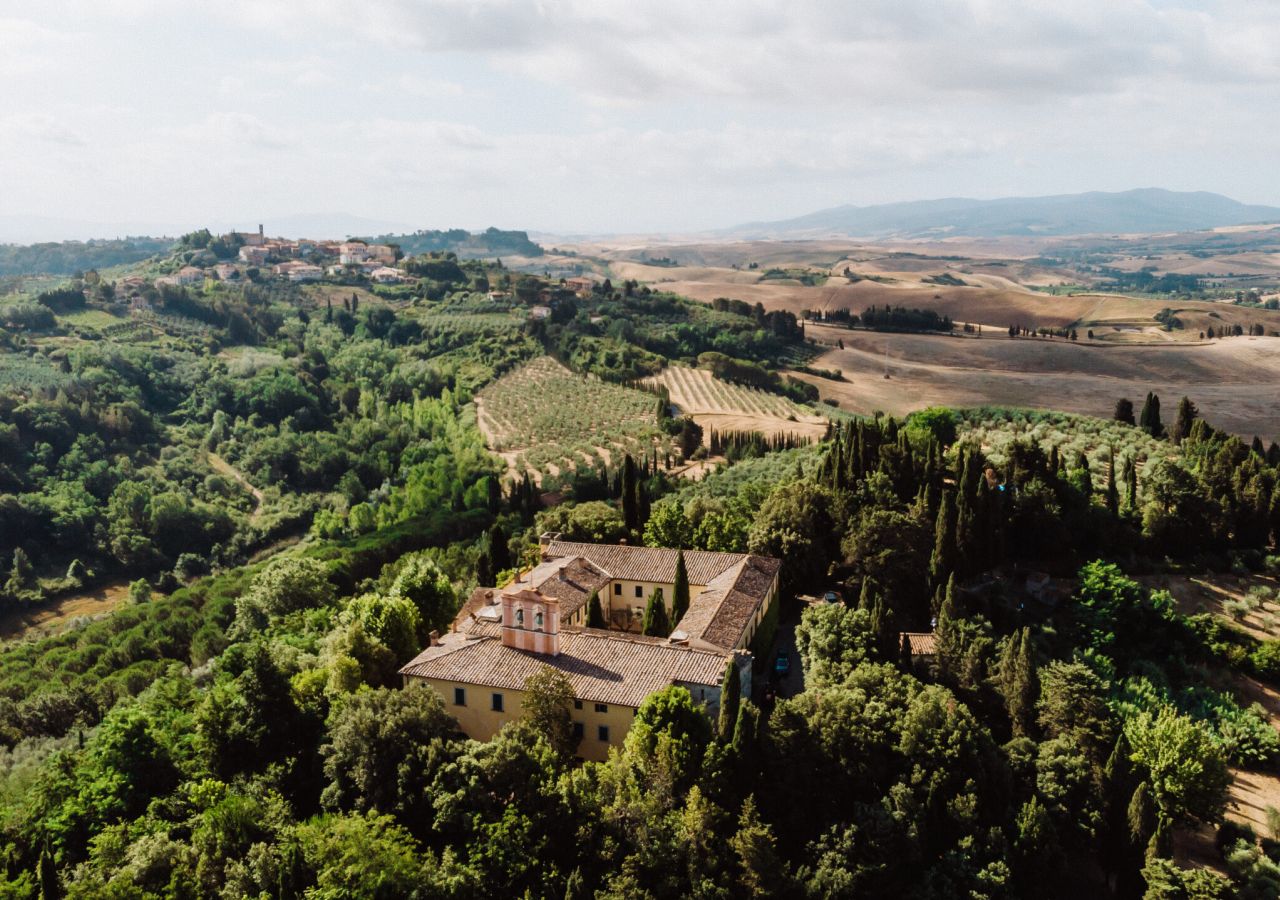 Veduta aerea di Badia di Morrona, immersa nel verde, circondata da cipressi, uliveti e campi coltivati, con un borgo e le colline toscane sullo sfondo.