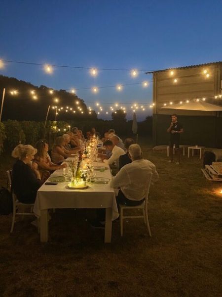 Cena serale tra i filari della Tenuta La Macchia, con numerosi ospiti seduti a una lunga tavola illuminata da candele e fili di luci sospese. Sullo sfondo, una persona in piedi si rivolge ai partecipanti.