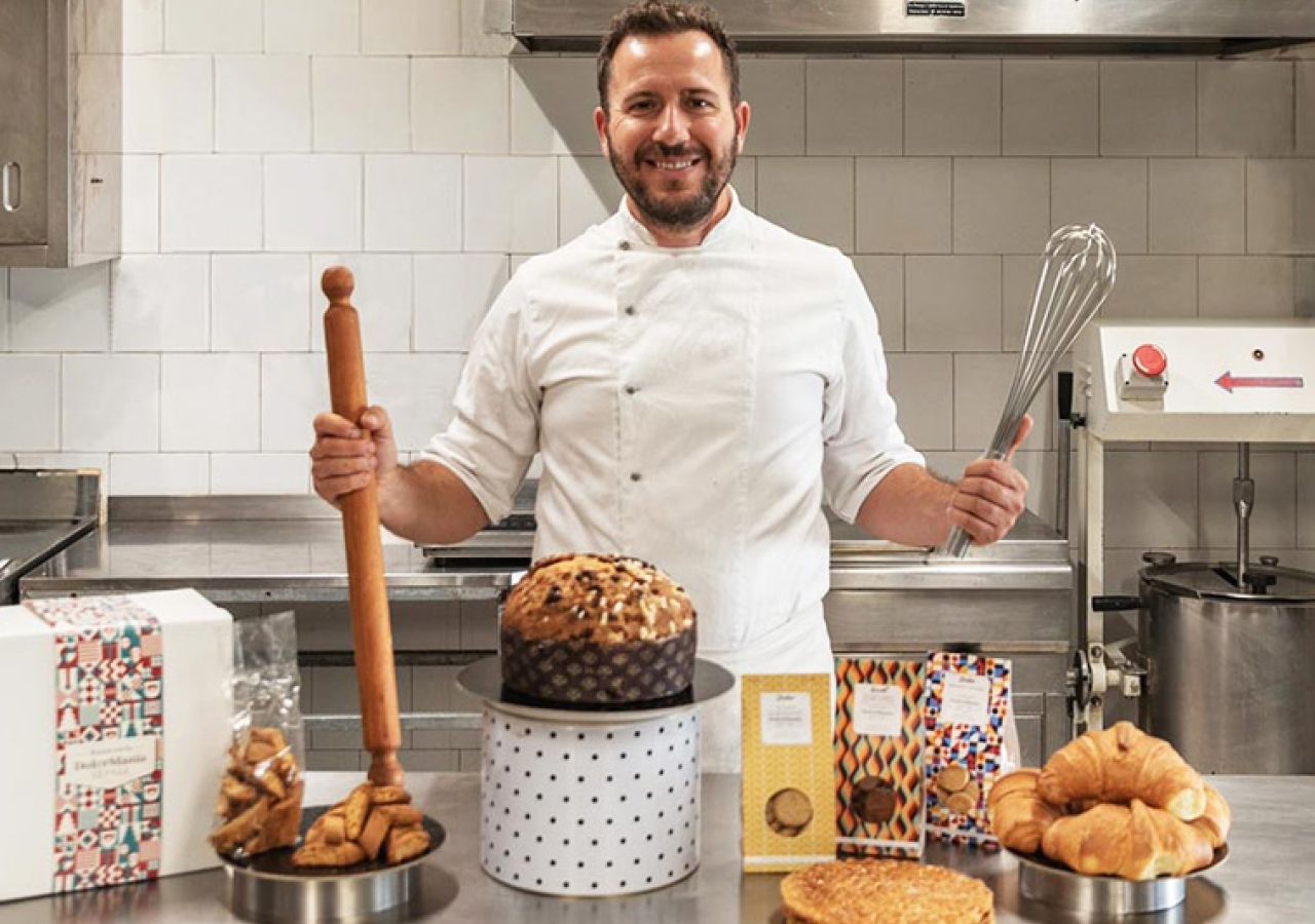 Pasticcere nel laboratorio di DolceMania Italia, circondato da panettone, biscotti, croissant e altri prodotti di pasticceria artigianale.