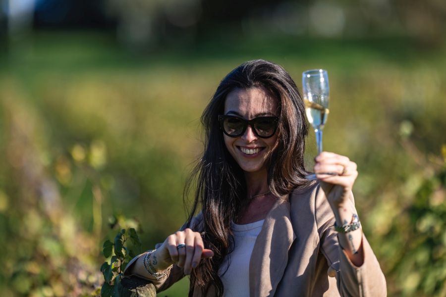 Donna sorridente tra i filari della tenuta L’Agona mentre solleva un calice di vino bianco.