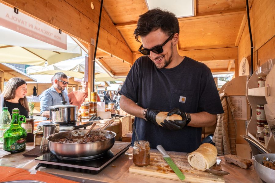 Cuoco durante una dimostrazione gastronomica all’aperto, mentre prepara e confeziona un ragù in barattolo davanti a una padella, con pane e ingredienti sul banco.