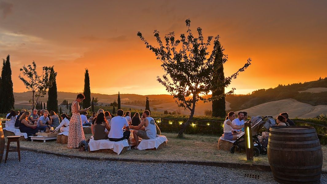 Persone sedute a tavola durante una cena all’aperto al tramonto, tra cipressi e colline toscane, con il cielo illuminato da calde tonalità arancioni.