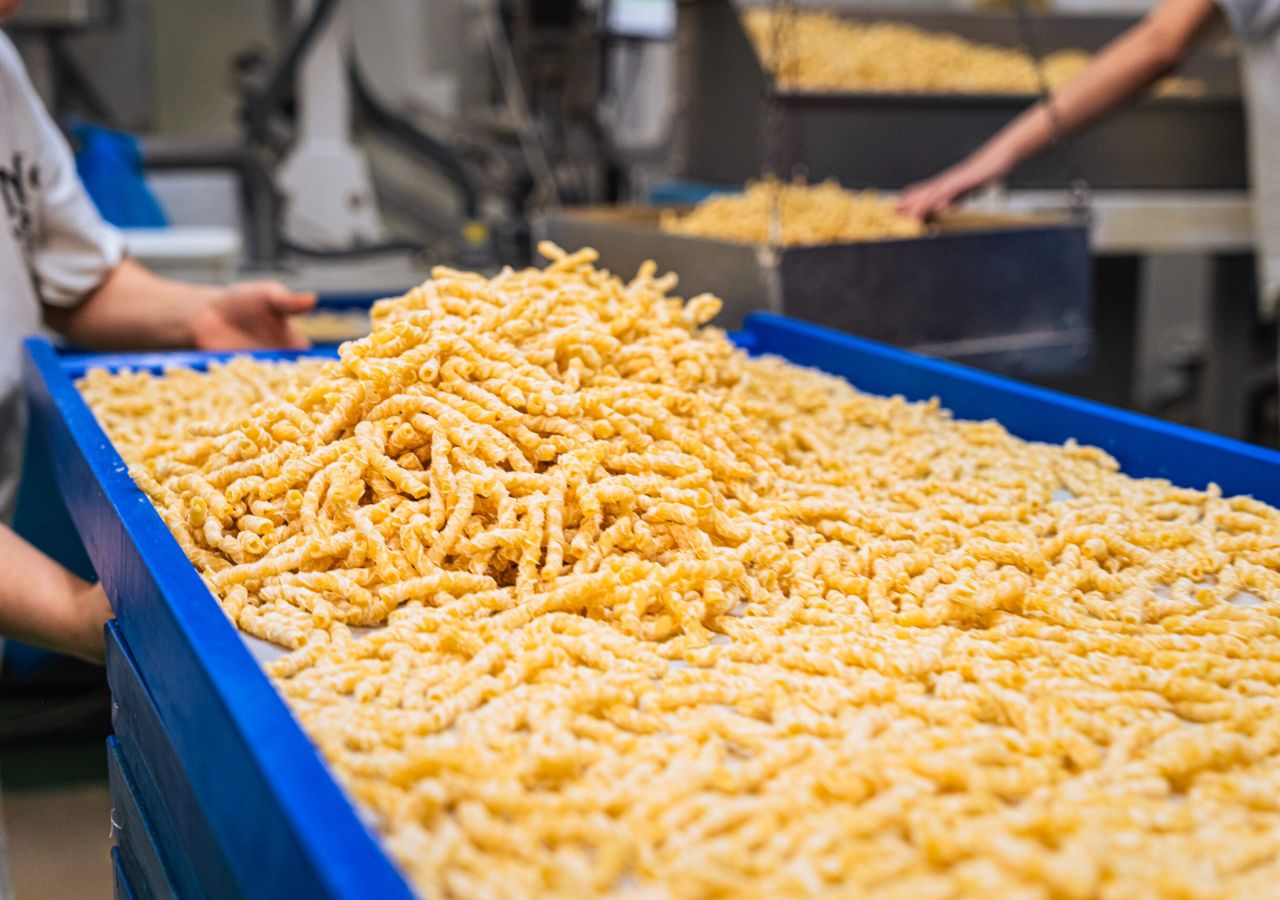 Pasta corta artigianale appena prodotta, raccolta in grandi contenitori blu all’interno del pastificio Morelli, con gli addetti al lavoro sullo sfondo.
