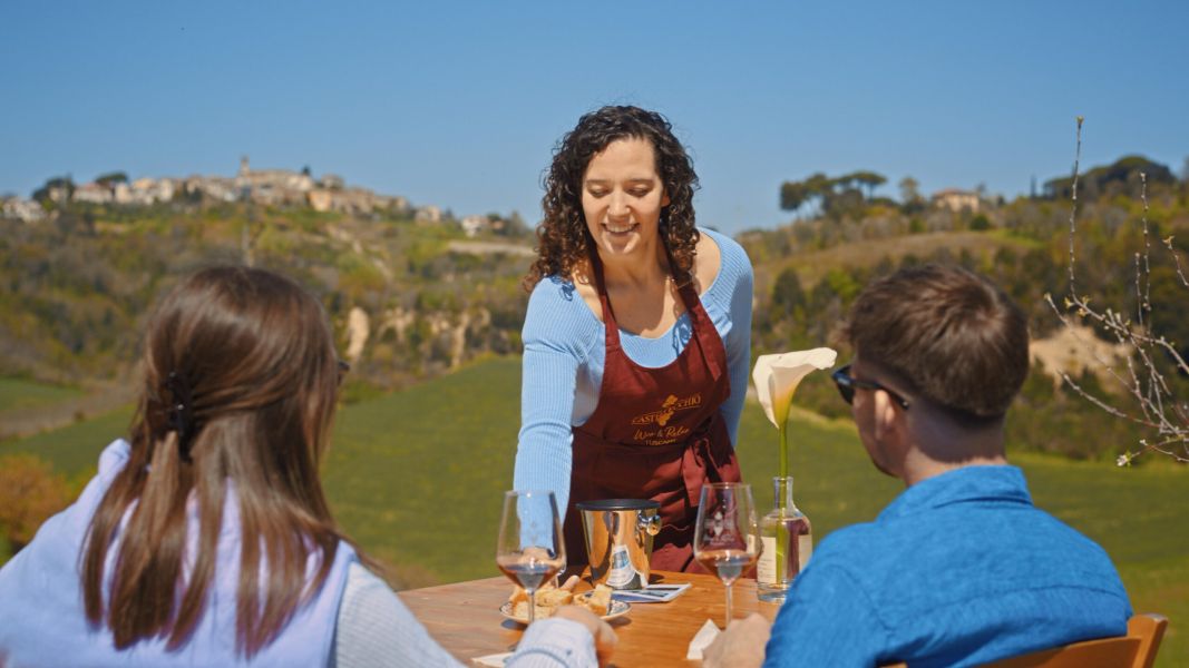 Una donna con grembiule di Castelvecchio serve una degustazione di vino a due ospiti seduti a un tavolo all’aperto, con le colline e un borgo sullo sfondo.