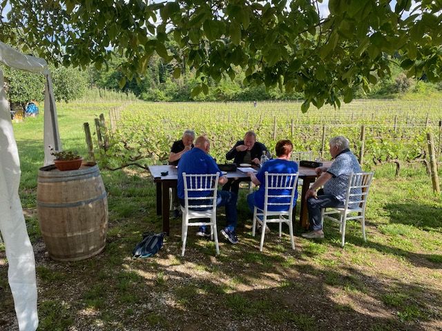 Cinque persone siedono attorno a un tavolo all’aperto, all’ombra di un albero, durante un momento conviviale tra i filari della Tenuta La Macchia. Sul lato sinistro si vede una botte in legno, mentre sullo sfondo si estende il vigneto.