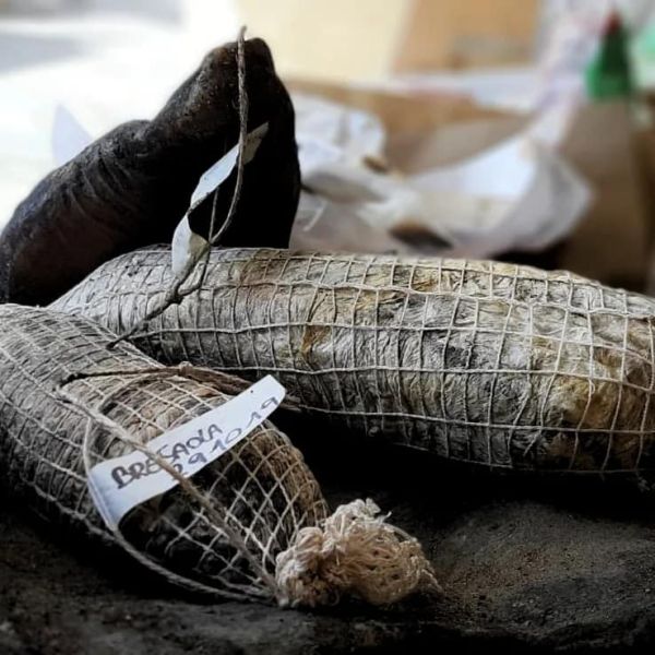 Salumi artigianali stagionati, avvolti in rete e legati con spago, disposti su un piano di lavoro con piccole etichette manoscritte.